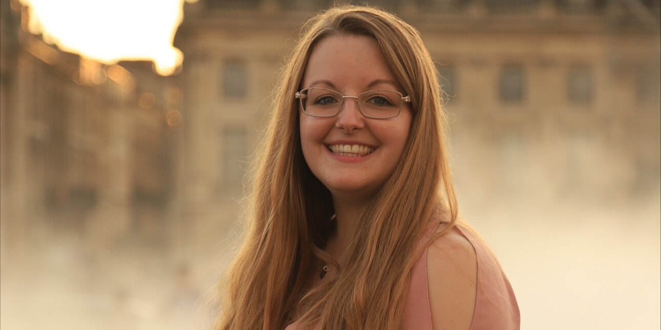 A portrait of a smiling Rose Ling, a white woman with long hair and glasses, stood side on in front of old yellow brick buildings
